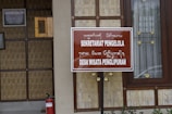 A wooden and bamboo structure with a brown sign displaying text in both a foreign script and in Latin script reading 'SEKRETARIAT PENGELOLA DESA WISATA PENGLIPURAN'. The background includes a framed certificate on the wall and a window with decorative elements. A red fire extinguisher is placed near the bottom left.