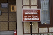 A wooden and bamboo structure with a brown sign displaying text in both a foreign script and in Latin script reading 'SEKRETARIAT PENGELOLA DESA WISATA PENGLIPURAN'. The background includes a framed certificate on the wall and a window with decorative elements. A red fire extinguisher is placed near the bottom left.