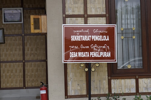 A wooden and bamboo structure with a brown sign displaying text in both a foreign script and in Latin script reading 'SEKRETARIAT PENGELOLA DESA WISATA PENGLIPURAN'. The background includes a framed certificate on the wall and a window with decorative elements. A red fire extinguisher is placed near the bottom left.