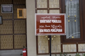 A wooden and bamboo structure with a brown sign displaying text in both a foreign script and in Latin script reading 'SEKRETARIAT PENGELOLA DESA WISATA PENGLIPURAN'. The background includes a framed certificate on the wall and a window with decorative elements. A red fire extinguisher is placed near the bottom left.