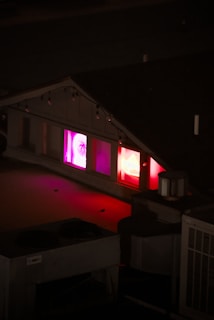 A dimly lit rooftop scene at night with neon lights visible through the windows of a small building. The neon signs glow in shades of pink and red, creating a vibrant contrast with the dark surroundings.