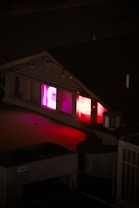 A dimly lit rooftop scene at night with neon lights visible through the windows of a small building. The neon signs glow in shades of pink and red, creating a vibrant contrast with the dark surroundings.