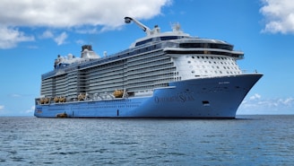 A large cruise ship sails on a calm sea under a clear blue sky. The ship, called Ovation of the Seas, has multiple decks with rows of windows and lifeboats. The ocean reflects the blue of the sky, creating a serene scene.