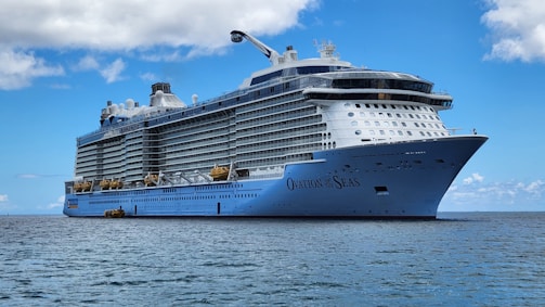 A large cruise ship sails on a calm sea under a clear blue sky. The ship, called Ovation of the Seas, has multiple decks with rows of windows and lifeboats. The ocean reflects the blue of the sky, creating a serene scene.