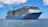 A large cruise ship sails on a calm sea under a clear blue sky. The ship, called Ovation of the Seas, has multiple decks with rows of windows and lifeboats. The ocean reflects the blue of the sky, creating a serene scene.