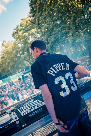 A person wearing a sports jersey with the name 'Pippen' and the number '33' is standing at a DJ booth, overseeing a large crowd gathered outdoors. Surrounding the DJ setup are mixing equipment and turntables. Bright sunlight filters through the trees, suggesting a lively outdoor event.