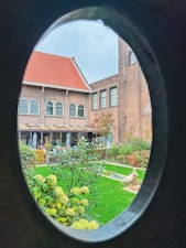A picturesque garden view is framed by an oval window or opening, showcasing a well-maintained lawn, various flowering plants, and shrubs. The background features a charming brick building with arched windows and a red-tiled roof, creating an inviting and serene atmosphere.