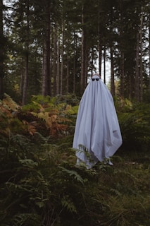 A figure resembling a ghost, covered entirely by a white sheet with sunglasses, stands amidst a dense forest. The background is composed of tall trees and lush green ferns in a natural setting.