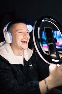 A smiling person wearing headphones is recording themselves with a smartphone placed on a ring light stand. The background is dimly lit, focusing attention on the subject who appears to be engaged with the camera, possibly live streaming or recording a video.