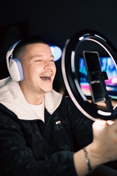 A smiling person wearing headphones is recording themselves with a smartphone placed on a ring light stand. The background is dimly lit, focusing attention on the subject who appears to be engaged with the camera, possibly live streaming or recording a video.
