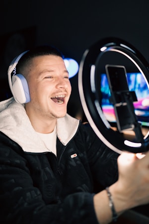 A smiling person wearing headphones is recording themselves with a smartphone placed on a ring light stand. The background is dimly lit, focusing attention on the subject who appears to be engaged with the camera, possibly live streaming or recording a video.