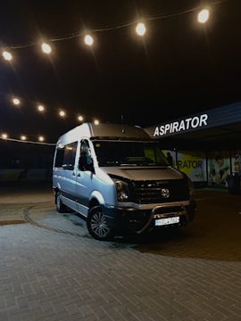 A silver van parked under string lights at night, with a sign that reads 'ASPIRATOR' in the background. The area is paved with bricks and the lights create a warm glow.