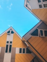Distinctive yellow and grey wooden cube-like structures with slanted roofs and multiple windows are set against a bright blue sky.