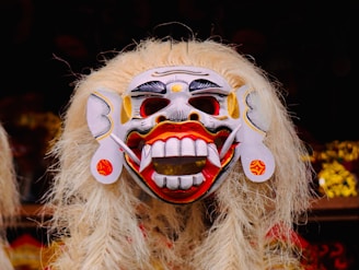 An ornate traditional mask with exaggerated features and long, flowing white hair. The mask is primarily white with striking red and black accents, featuring large eyes, prominent teeth, and decorative elements around the ears.