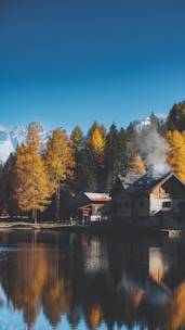 A serene mountain lake scene with a rustic house on the shore. Smoke rises from a chimney, indicating a fire burning inside. Tall trees with autumn foliage surround the lake, creating a mirror-like reflection on the water. Snow-capped mountains are visible in the background under a clear blue sky.