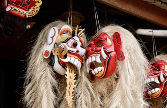 Elaborately carved wooden masks with intricate details and vibrant colors, featuring exaggerated facial features such as wide eyes and large teeth. Each mask has long, light-colored hair cascading down, and they are arranged closely together.
