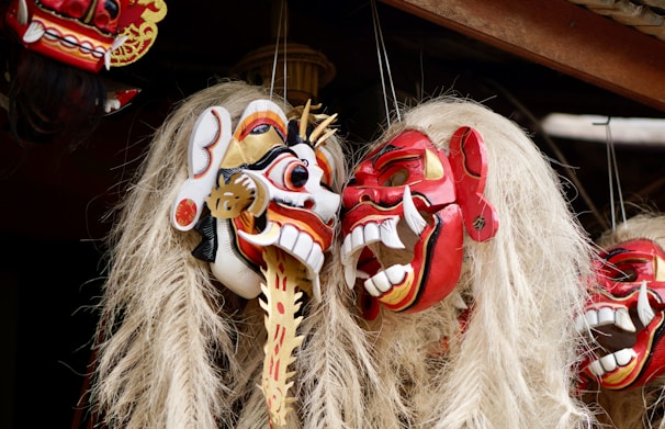 Elaborately carved wooden masks with intricate details and vibrant colors, featuring exaggerated facial features such as wide eyes and large teeth. Each mask has long, light-colored hair cascading down, and they are arranged closely together.