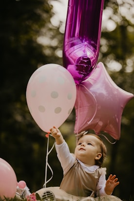A young child reaches up towards colorful balloons, including a shimmering pink star and a large numeral one, set against a blurred natural background.