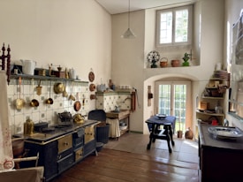 A vintage kitchen with a large black and brass stove, surrounded by white and green patterned tiles. Various copper and brass utensils hang on the wall. A small wooden table with a blue tablecloth sits near a window with white frames that allow natural light to fill the room. Shelves hold ceramic and metal pots, and there is a variety of kitchenware throughout the space.