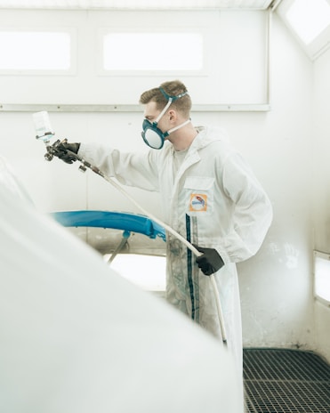 A person wearing protective gear including a mask and gloves is engaged in spray painting. The setting appears industrial, with white walls and a gridded floor. The individual is focused on painting an object, possibly a car part, blue.