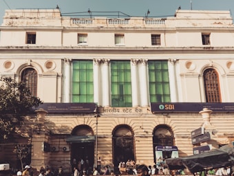 A historic building with classical architecture featuring tall, white columns and large, arched windows. The facade is adorned with a mix of cream-colored walls and green glass panels. A sign with 'SBI' indicates a bank located at street level. People are walking in front of the building, and there is a tree to the left casting shadows on the structure.