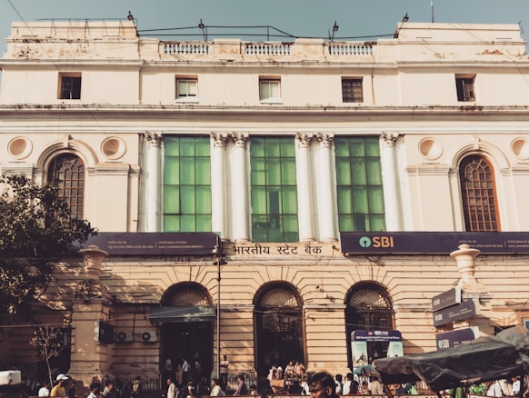 A historic building with classical architecture featuring tall, white columns and large, arched windows. The facade is adorned with a mix of cream-colored walls and green glass panels. A sign with 'SBI' indicates a bank located at street level. People are walking in front of the building, and there is a tree to the left casting shadows on the structure.