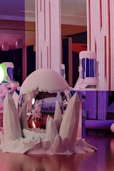 A futuristic interior space with large vertical light panels and colorful ambient lighting. The setup includes a spherical metallic object with a reflective surface surrounded by crystalline structures. A humanoid figure is visible near the object, contributing to a sci-fi theme.