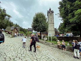 People are gathered in a cobblestone courtyard near a tall stone clock tower surrounded by lush green trees. Families and visitors, some with dogs, are walking or sitting on the stone wall, with a small wooden chapel visible in the background.