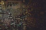 A chaotic chalkboard filled with various symbols, numbers, and text related to hacking. Words like 'HACKER', 'ATTACK', and 'UPLOAD' are prominently visible amidst a jumble of codes and cryptic symbols. The board conveys an atmosphere of mystery and complexity, reminiscent of a hacker's workspace.