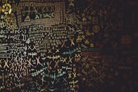 A chaotic chalkboard filled with various symbols, numbers, and text related to hacking. Words like 'HACKER', 'ATTACK', and 'UPLOAD' are prominently visible amidst a jumble of codes and cryptic symbols. The board conveys an atmosphere of mystery and complexity, reminiscent of a hacker's workspace.