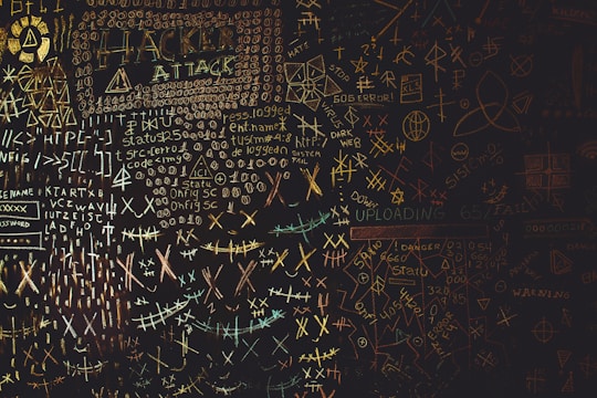 A chaotic chalkboard filled with various symbols, numbers, and text related to hacking. Words like 'HACKER', 'ATTACK', and 'UPLOAD' are prominently visible amidst a jumble of codes and cryptic symbols. The board conveys an atmosphere of mystery and complexity, reminiscent of a hacker's workspace.