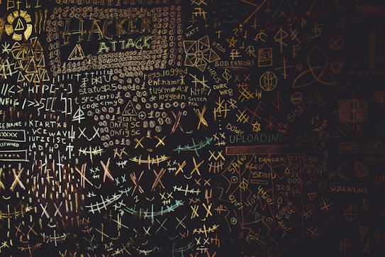 A chaotic chalkboard filled with various symbols, numbers, and text related to hacking. Words like 'HACKER', 'ATTACK', and 'UPLOAD' are prominently visible amidst a jumble of codes and cryptic symbols. The board conveys an atmosphere of mystery and complexity, reminiscent of a hacker's workspace.