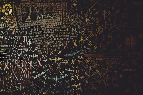 A chaotic chalkboard filled with various symbols, numbers, and text related to hacking. Words like 'HACKER', 'ATTACK', and 'UPLOAD' are prominently visible amidst a jumble of codes and cryptic symbols. The board conveys an atmosphere of mystery and complexity, reminiscent of a hacker's workspace.