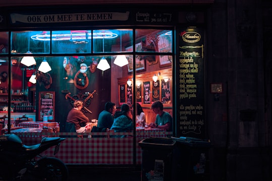 A small group of people sit inside a cozy restaurant with checkered tablecloths and dim lighting. The interior is adorned with vintage decor, including framed pictures and animal heads mounted on the wall. The neon sign and menu display add a quaint charm to the scene viewed from outside through the large window.