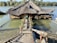 A wooden house with a thatched roof is situated on a body of water, accessible by a wooden footbridge. The structure is adorned with plants and flowers on either side. Two life buoys are visible, and a person is walking on the bridge towards the house. In the background, there is a lush, tree-lined shore.
