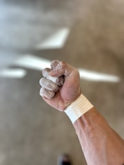 A close-up view of a clenched fist with chalk-covered fingers and wrist supported by athletic tape. The background is blurred, highlighting the focus on the hand.