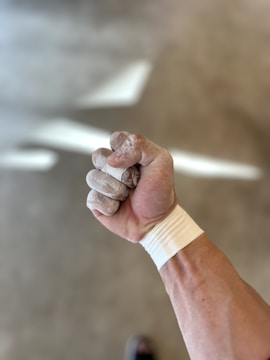 A close-up view of a clenched fist with chalk-covered fingers and wrist supported by athletic tape. The background is blurred, highlighting the focus on the hand.