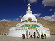 a white building with a gold roof and a gold top with people walking around with Shanti Stupa in the background