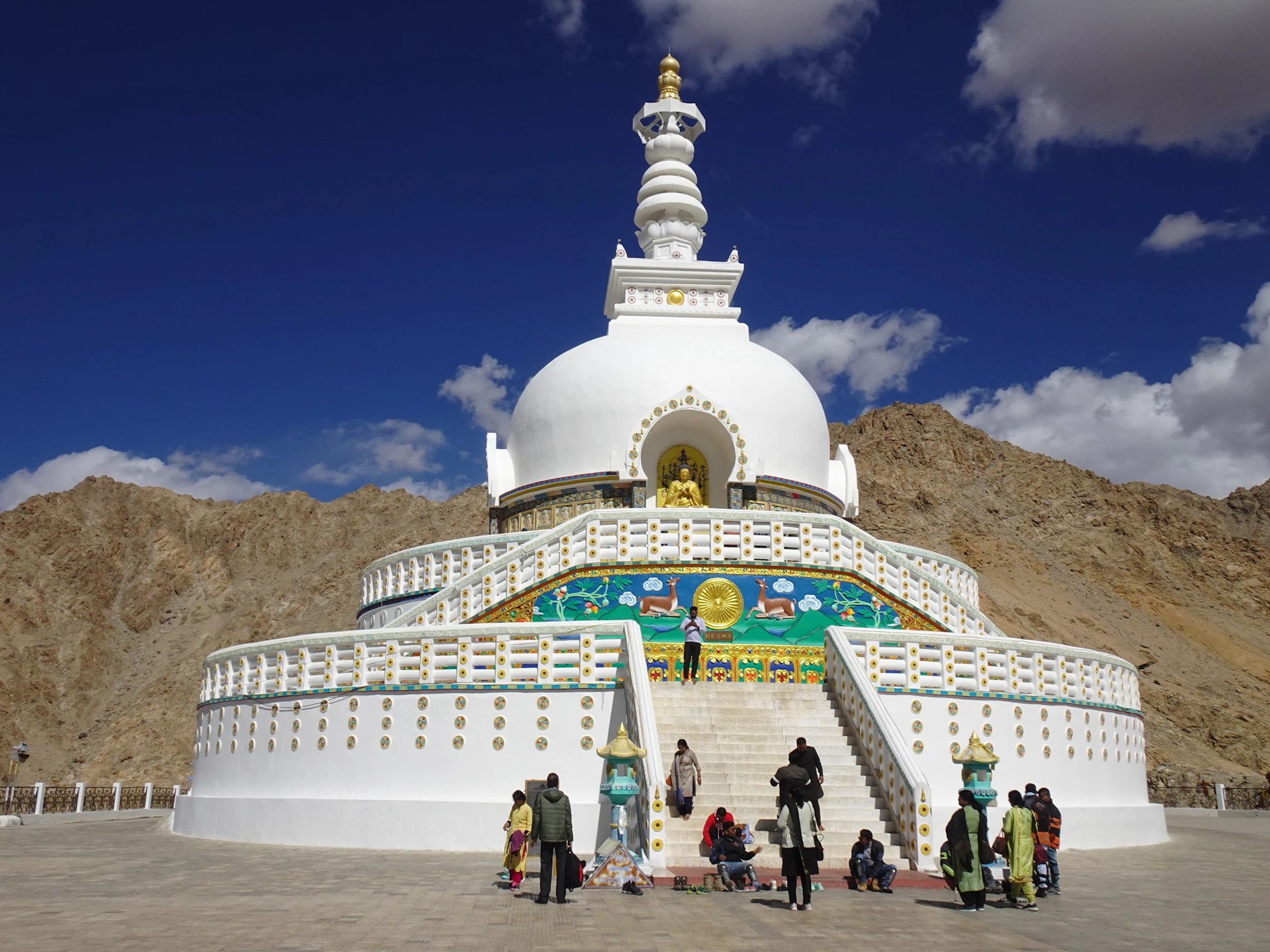 a white building with a gold roof and a gold top with people walking around with Shanti Stupa in the background