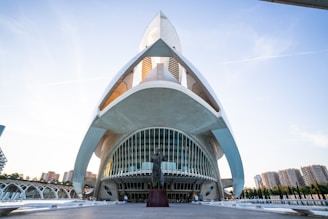 A futuristic architectural structure with sweeping, curved lines and a prominent archway. The building has large, reflective glass panels and white concrete surfaces. In the foreground, there is a statue on a pedestal. The background shows a clear blue sky and a few high-rise buildings.