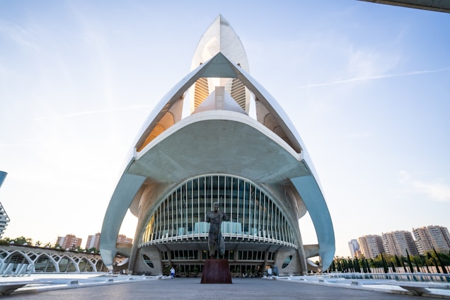 A futuristic architectural structure with sweeping, curved lines and a prominent archway. The building has large, reflective glass panels and white concrete surfaces. In the foreground, there is a statue on a pedestal. The background shows a clear blue sky and a few high-rise buildings.