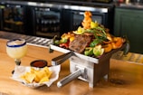 A rustic wooden table displays an enticing array of food, featuring a metal container filled with grilled meats, vegetables, and lime wedges. The colorful presentation includes green peppers, lemons, and onions. To the left, a frosty drink with a salted rim and a slice of lime sits next to a small dish of sauce and a bowl of tortilla chips.