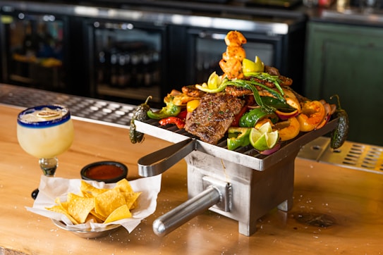 A rustic wooden table displays an enticing array of food, featuring a metal container filled with grilled meats, vegetables, and lime wedges. The colorful presentation includes green peppers, lemons, and onions. To the left, a frosty drink with a salted rim and a slice of lime sits next to a small dish of sauce and a bowl of tortilla chips.