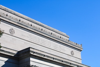 A classical architectural structure with decorative engravings and inscriptions. The building features intricate carvings near the roofline and an embossed medallion. The inscription reads, 'THE GLORY AND ROMANCE OF OUR HISTORY ARE HERE PRESERVED IN THE CHRONICLES OF THOSE WHO CONCEIVED AND BUILDED THE STRUCTURE OF OUR NATION.' A clear blue sky forms the backdrop.