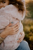 A person with light blue painted nails is holding a child dressed in a fluffy, light pink coat. The child's face is partially visible, nestled against the person's chest, creating a sense of warmth and protection. Sunlight softly illuminates the scene, highlighting the child's curly hair, and the background appears to be a meadow with tall grass.
