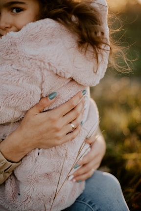A person with light blue painted nails is holding a child dressed in a fluffy, light pink coat. The child's face is partially visible, nestled against the person's chest, creating a sense of warmth and protection. Sunlight softly illuminates the scene, highlighting the child's curly hair, and the background appears to be a meadow with tall grass.