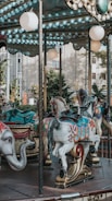 A carousel with elaborately decorated horses and an elephant figure. The carousel features bright colors and intricate designs on the horses. The background shows trees and part of a building, indicating an outdoor setting.