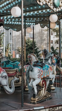 A carousel with elaborately decorated horses and an elephant figure. The carousel features bright colors and intricate designs on the horses. The background shows trees and part of a building, indicating an outdoor setting.