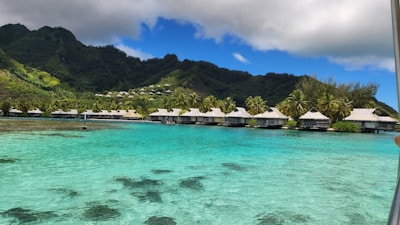 Overwater bungalows line a tranquil lagoon with crystal-clear turquoise water, surrounded by lush green mountains under a partly cloudy sky. Palm trees dot the shoreline, adding to the tropical setting.