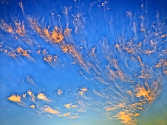 Wispy, orange-tinted clouds stretch across a deep blue sky, creating a dynamic and lively pattern. The clouds appear to be illuminated by the setting or rising sun, enhancing their golden hues.